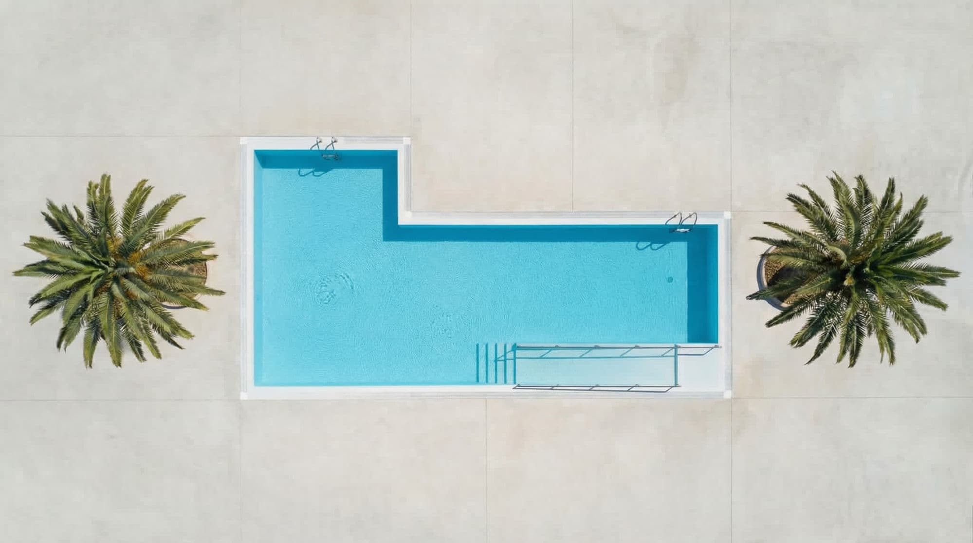 Vue aérienne drone de la piscine en L de la Maison Bledi, encadrée de deux palmiers dattiers et d'une rampe d'accès PMR en pente douce qui descend dans l'eau